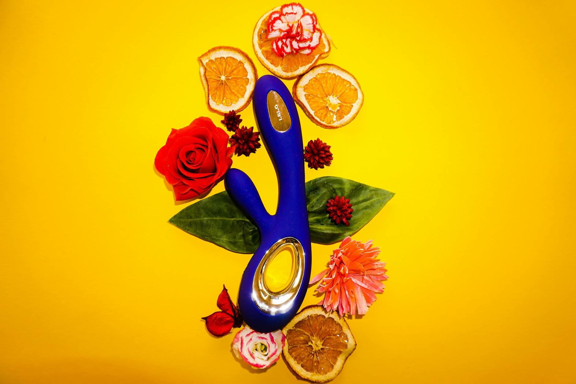 A blue lemon clitoral vibrator surrounded by fresh citrus and flowers on a bright yellow background
